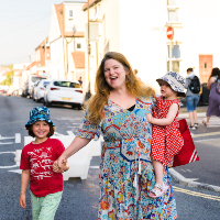 A mother and two young children walk down a residential street in Bristol. One holds her hand, the other sits on her hip. All are smiling, the day is bright and the weather looks warm.