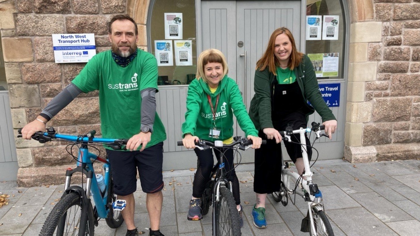 A man and two women stand with bikes outside an active travel centre wearing green Sustrans branded clothing.