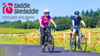 Two happy cyclists on a traffic-free cycle path surrounded by flowers. The sky is blue and the sun is shining. Saddle Skedaddle logo overlaid top left