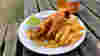 A plate of fish and chips with mushy peas on a wooden bench outside next to a bottle of beer