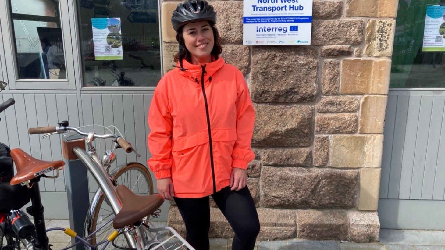 A woman wearing an orange jacket and black leggings and helmet stands beside bikes out an Active Travel Centre.