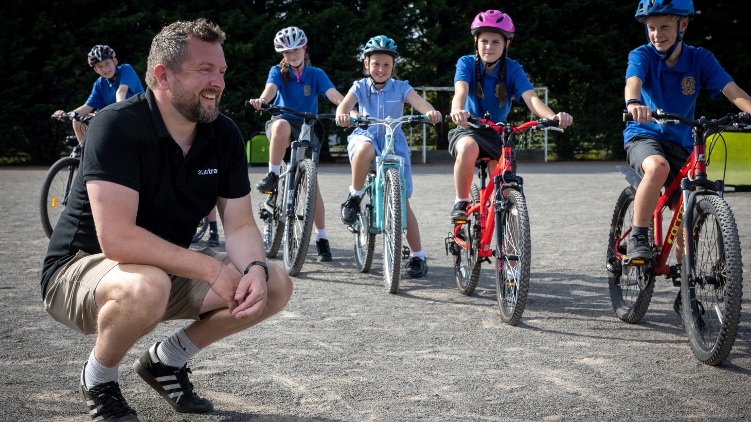 A male Sustrans active travel officer crouches on the ground in school playground while a row of children on bikes wearing helmets line up beside him