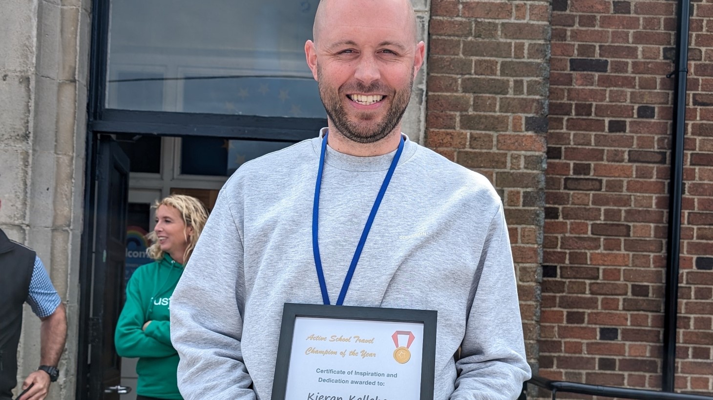 A man wearing a grey sweatshirt holds up a framed certificate while standing outside a school.