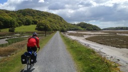 Cyclist in red jacket on gravel path by estuary with green hills and trees to the side